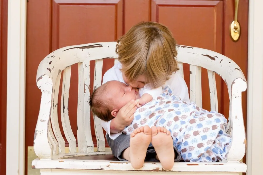 Toddler sitting in white striped chair holding and kissing newborn baby